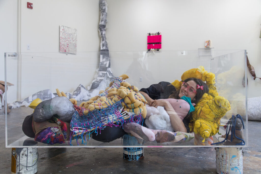 student laying in plexiglass box with soft sculptures