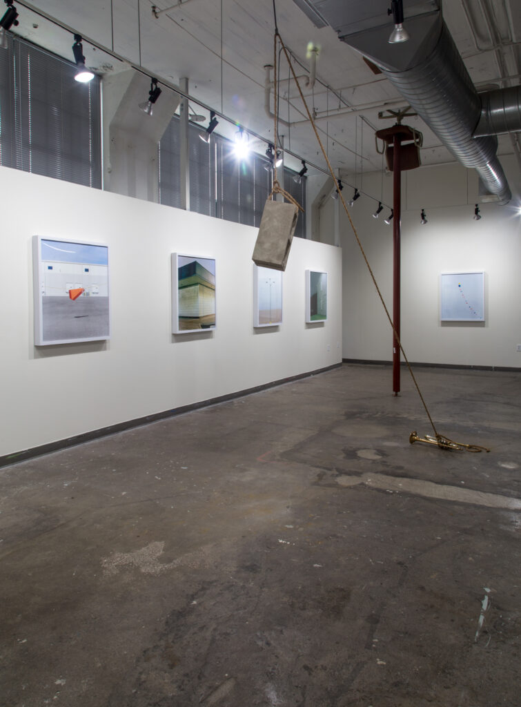 sculpture of cinderblock suspended from rope with other end attached to trumpet on floor with background of framed photographs