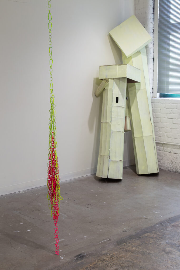 sculpture of hanging mass of bright green and pink keychains and rings in front of large painted cardboard sculpture