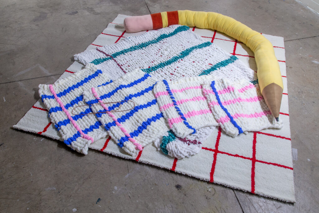 large textiles knitted to resemble lined paper with large soft sculpture of a number two pencil