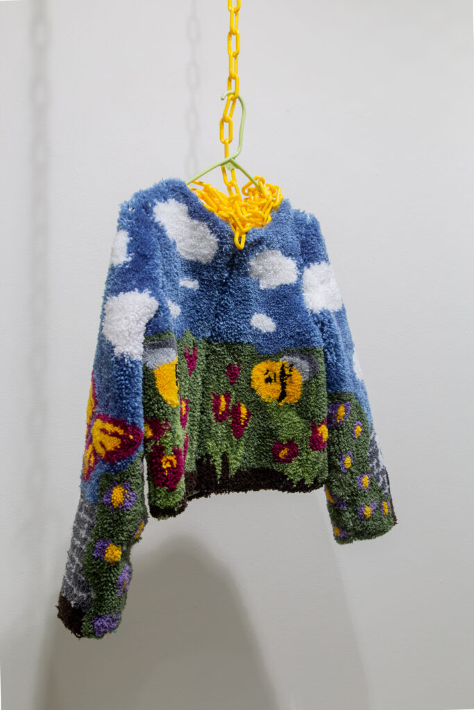 tufted yarn sweater depicting landscape with clouds and flowers suspended from yellow chain