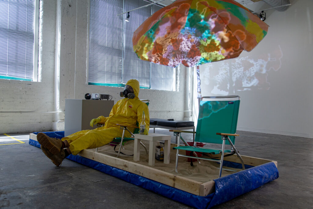 person in hazmat suit sitting on lawn chair in sandbox made of two-by-fours partially wrapped in blue tarp