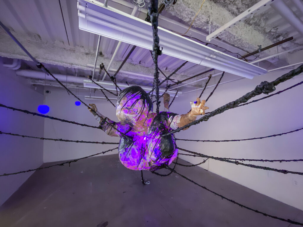 large sculpture of distorted figure suspended from black woven plastic ropes