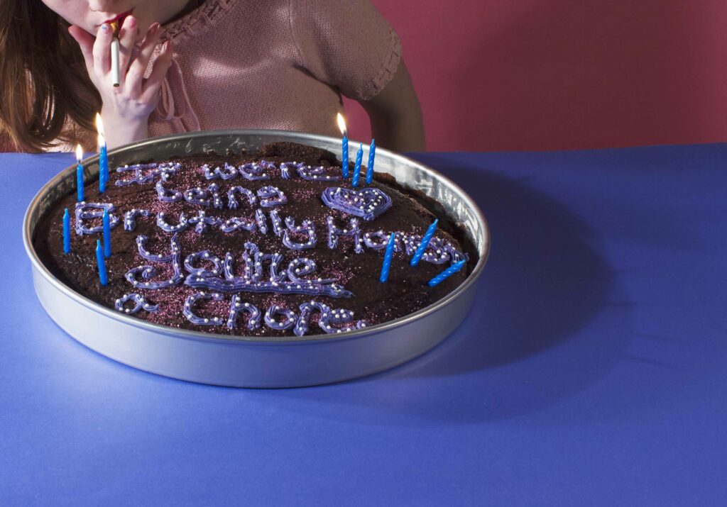 woman with unlit cigarette in mouth leaning over a cake with lit candles