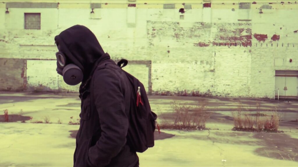 film still of person with gas mask and hoodie in urban landscape