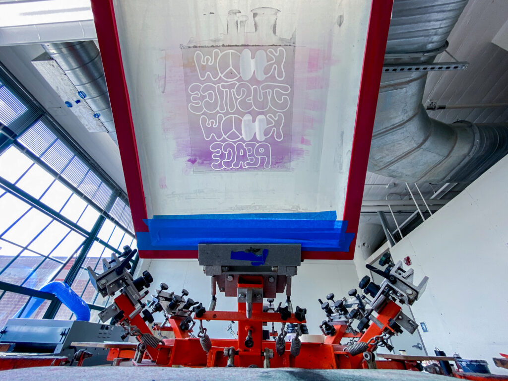 photo of a screen printing press with a screen reading "know justice know peace"