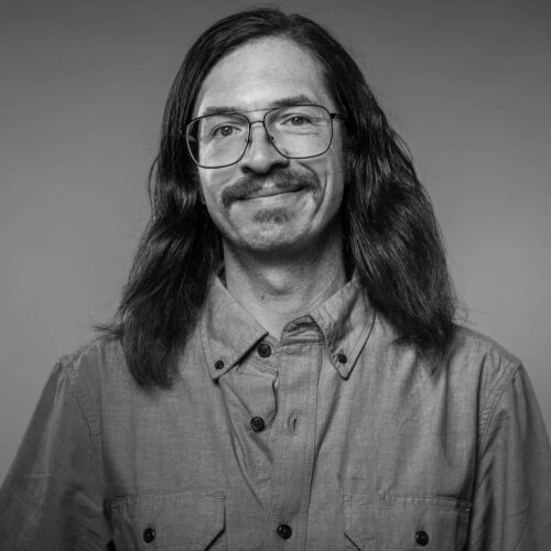 black and white headshot of Jimmy Baker (man smiling with glasses and long hair)