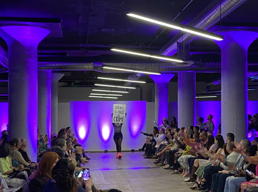 fashion show in site 1212 event space showing performer walking down runway with a sign reading "drag is not a crime"