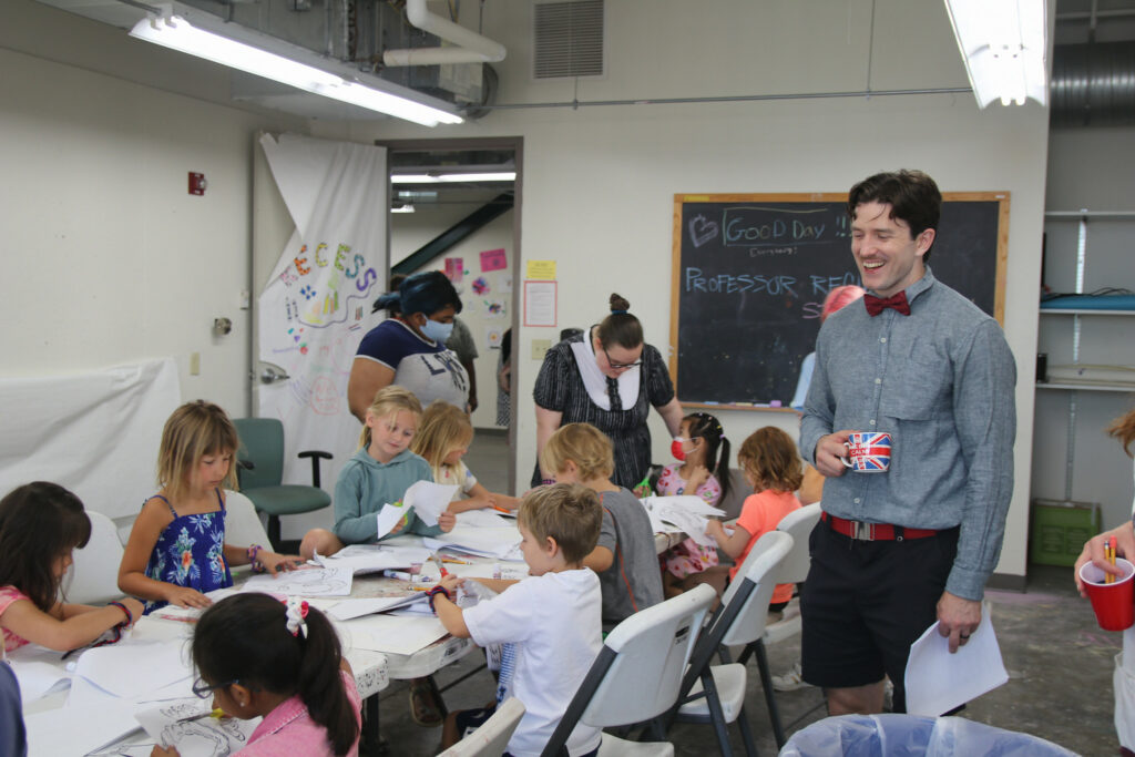 Three teachers assist nine students who make art at a table using paper, scissors, and pencils.