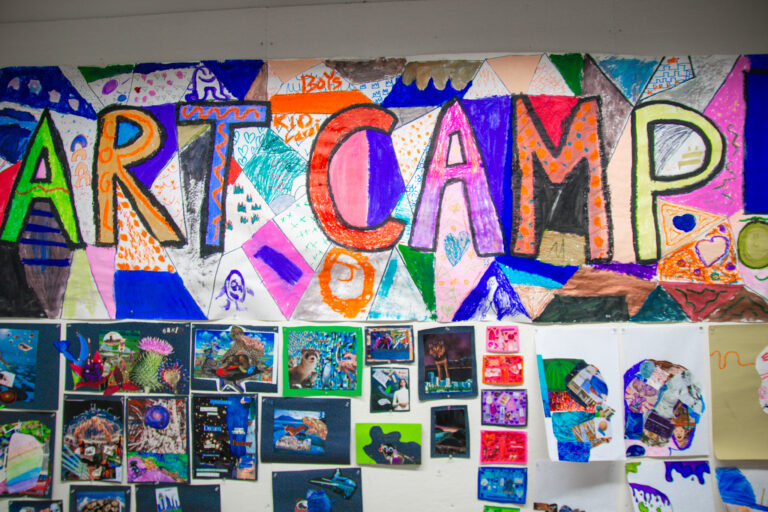 The words "Art Camp" are colorfully spelled out on a large banner with other 2D student artwork hung the the wall below it.