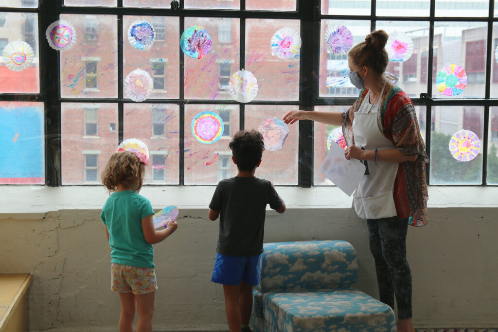 A teacher talks with two children about their artwork which is displayed on a large window.