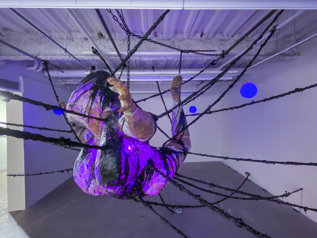 A self-illuminated sculpture of a rotund figure bound to the walls by many strands