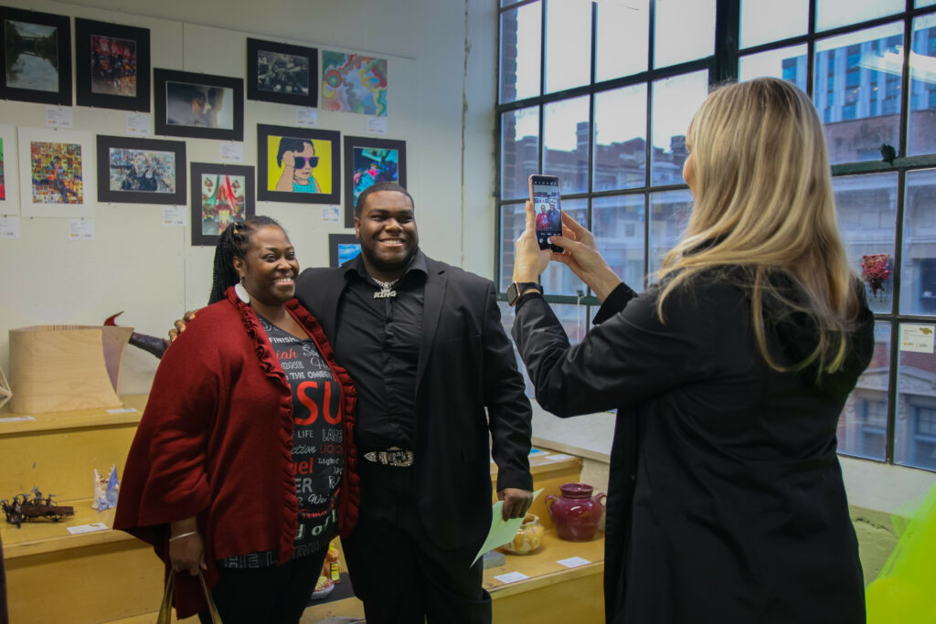 A woman photographing a student and an adult in front of Scholastic Works.