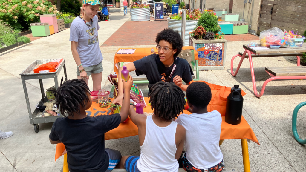 A young black man does arts and crafts with three black children at a picnic table in a park while a young white woman looks on