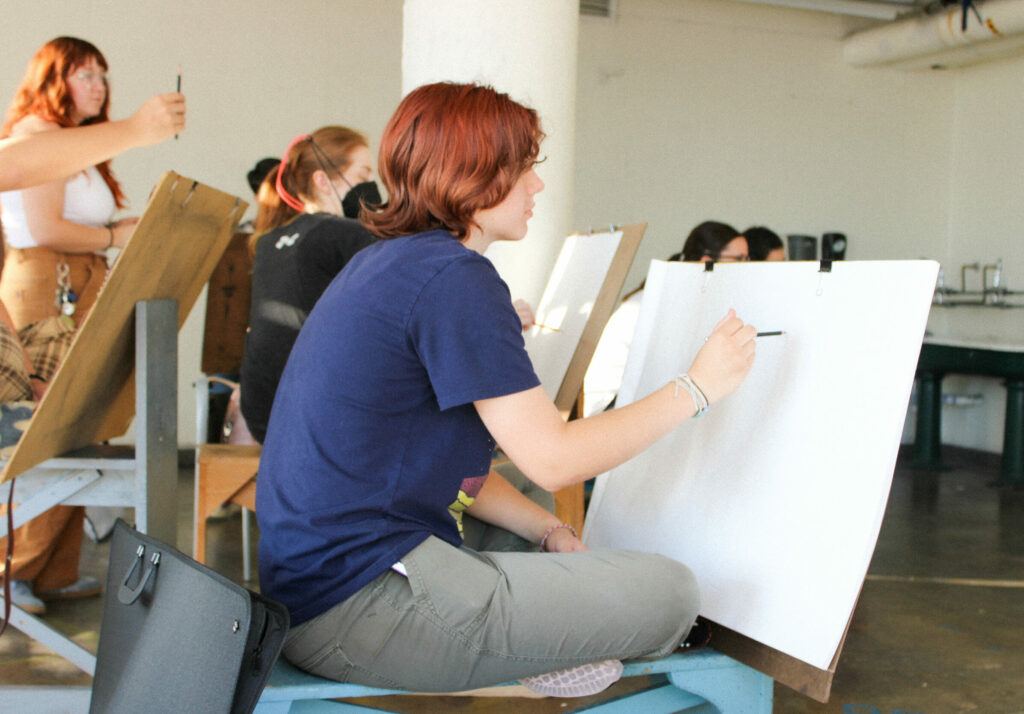 6 students draw a subject out of frame. One student is in focus. The instructor is in the background looking at the subject.