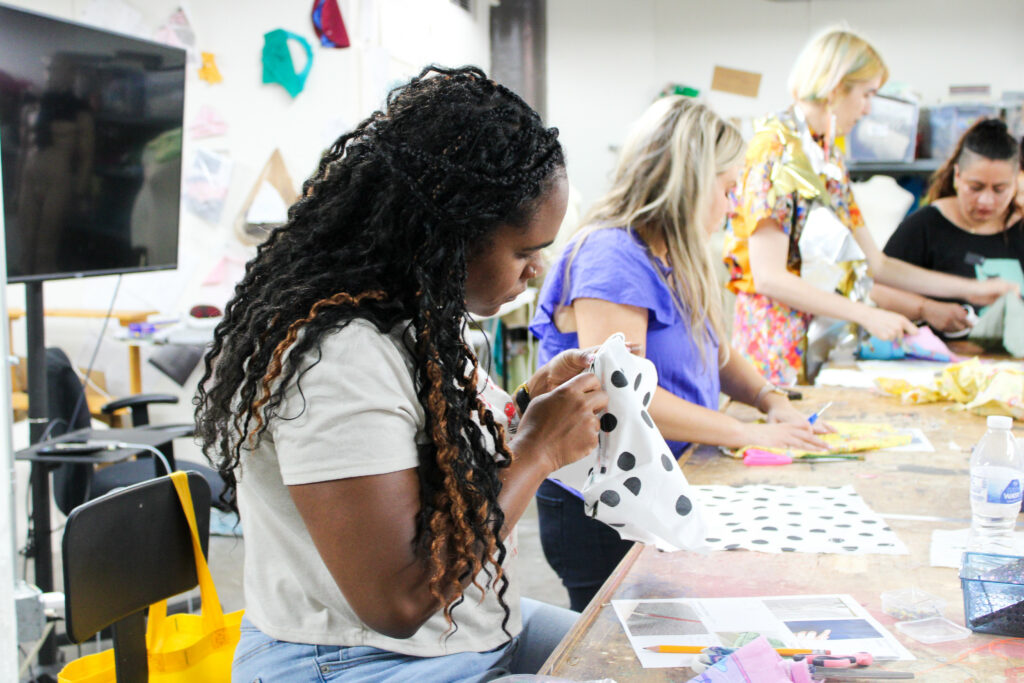 3 students work on sewing projects at a table while the instructor helps.