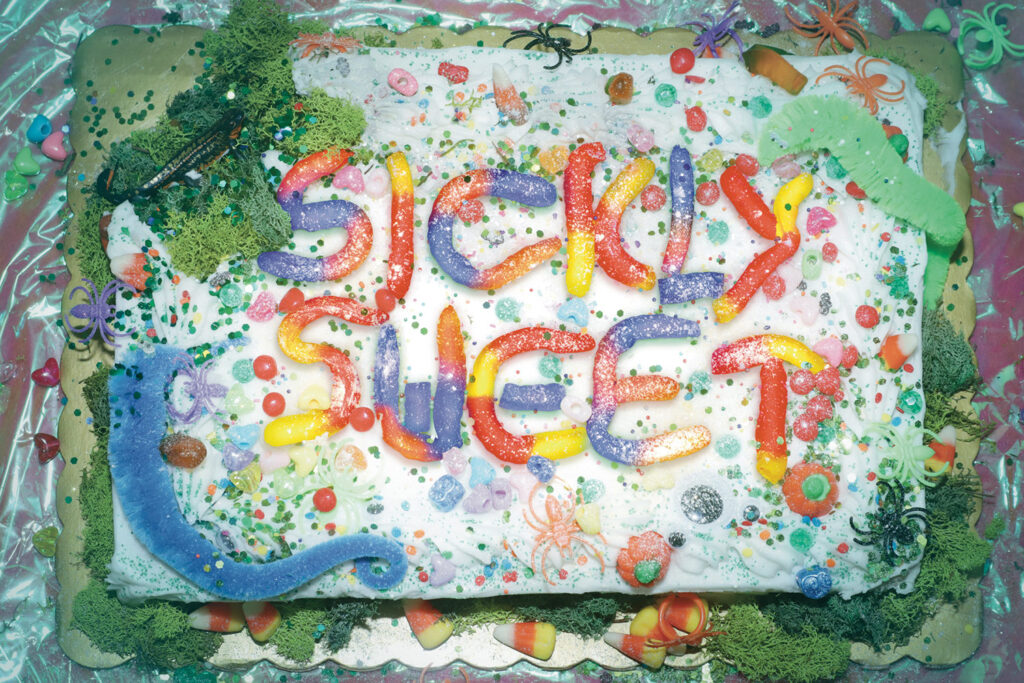 A photograph of a cake covered with various candy and objects that spell out the title "Sickly Sweet"