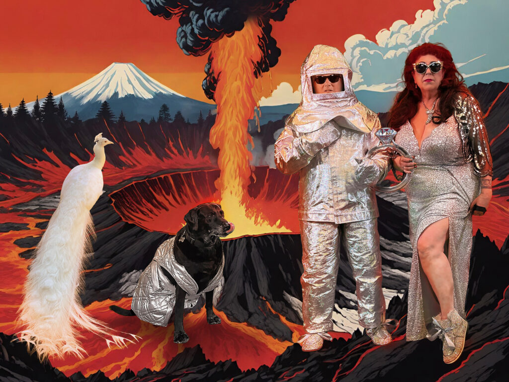 two women (Annie Sprinkle and Beth Stephens) in silver fire suits with a dog and bird imposed over a flaming volcano