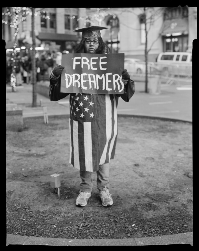 A graduate is standing with a sign that reads "Free Dreamers."