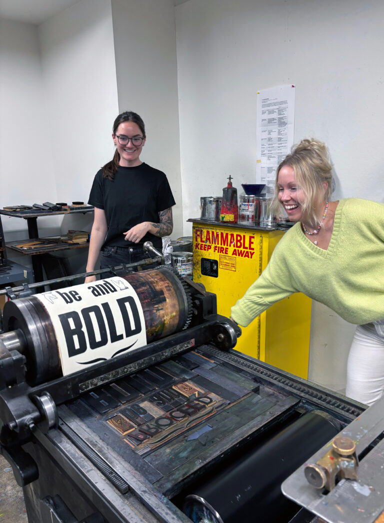 two women running a print through a letterpress machine that reads "Bold"