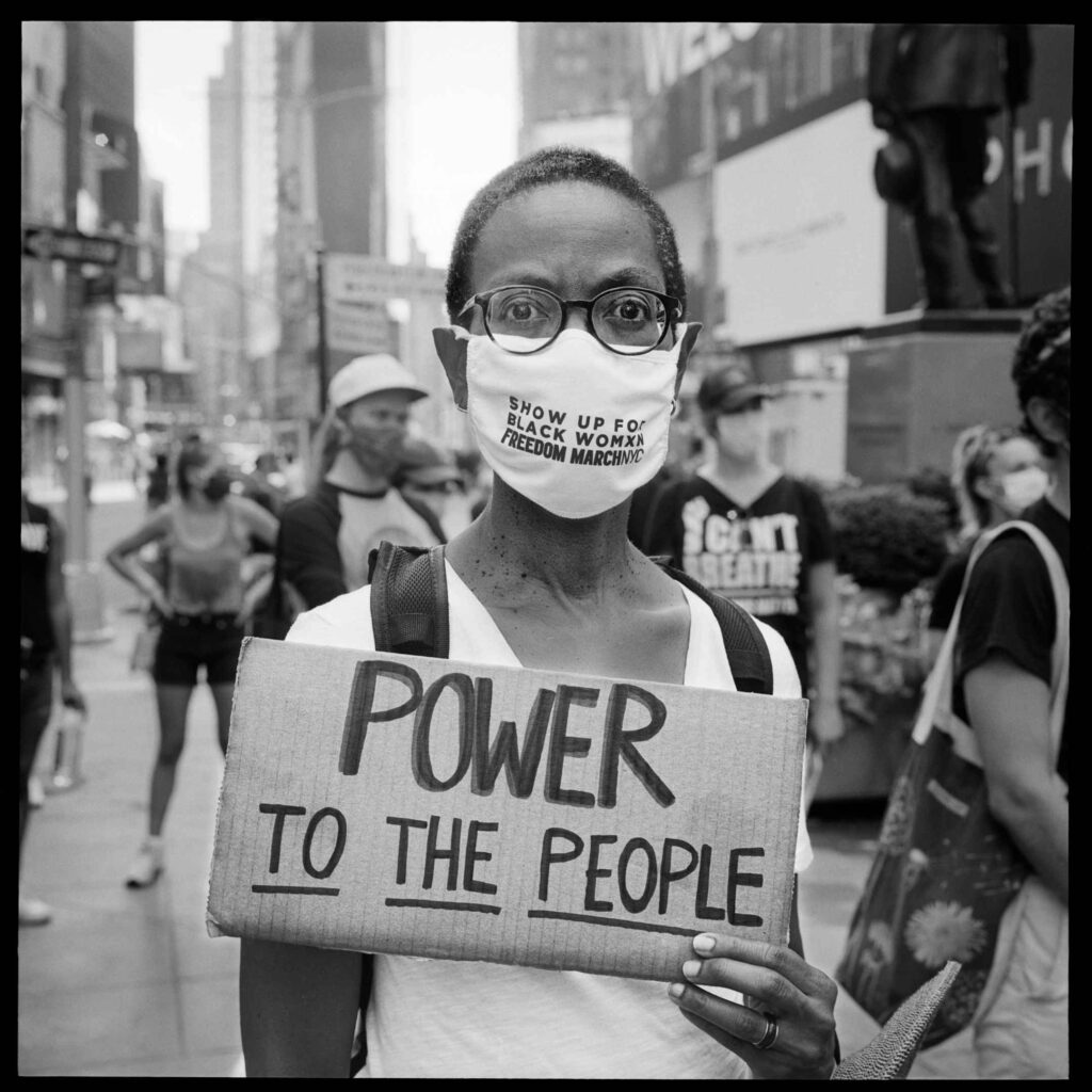 A woman in a mask holds a sign that reads "power to the people."